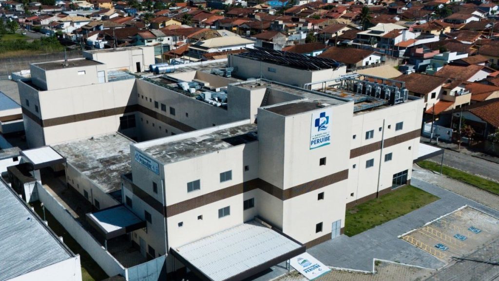 Vista aérea de um complexo de prédios industriais e residenciais com placa “PERUÍBE”, cercado por casas do bairro e áreas externas.