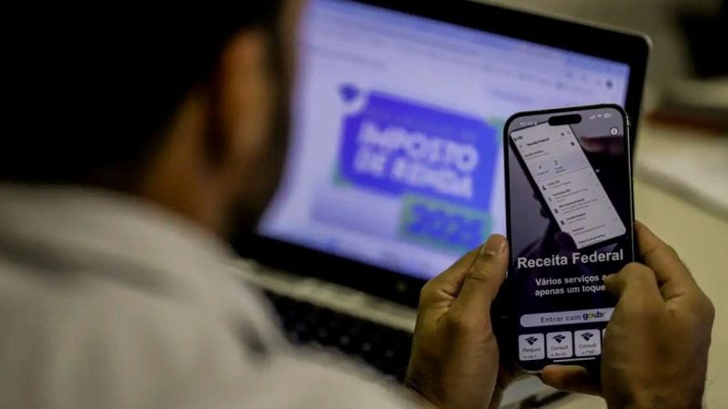 Pessoa usando celular com aplicativo da Receita Federal enquanto consulta informações de restituição do Imposto de Renda em um computador ao fundo
