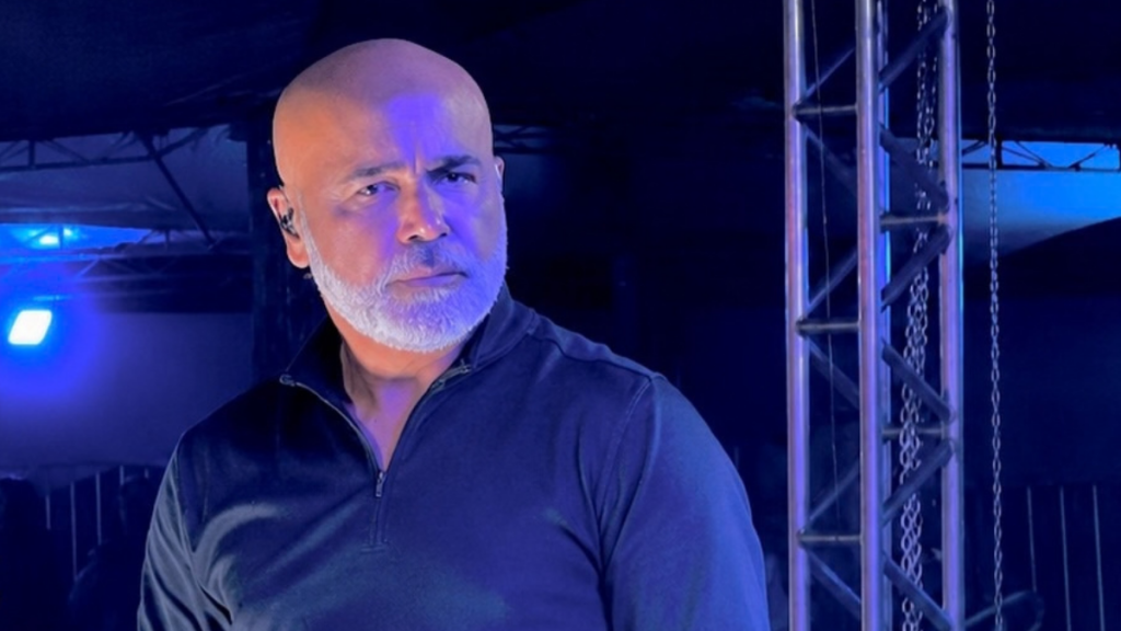 Texto alternativo (Alt Text):
O cantor sertanejo Rick (da dupla Rick & Renner), careca e de barba grisalha aparada, vestindo uma camisola preta de manga comprida com gola alta e fecho. Ele está num ambiente de palco ou bastidores com iluminação azul ao fundo e uma estrutura metálica à sua direita, olhando fixamente para o lado com uma expressão séria.