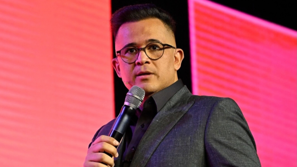 Bruno Avelar a falar ao microfone num palco, vestindo um fato cinzento e óculos de armação redonda, com um fundo iluminado em tom rosa.
