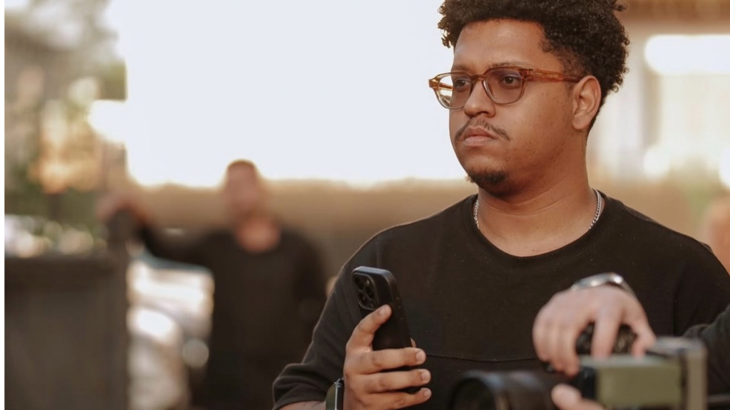 Fotografia de plano médio do videomaker Paulo Soares, um homem negro de óculos de armação clara e t-shirt preta, segurando um smartphone enquanto acompanha uma gravação de vídeo num ambiente exterior.