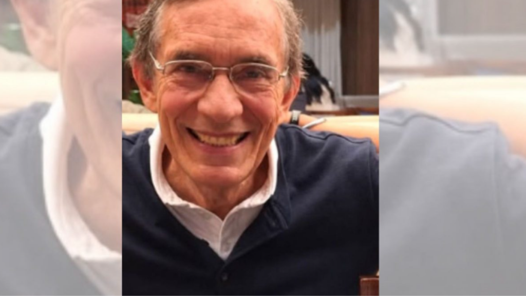 Fotografia em plano médio de Antonio Roberto Nóbrega de Araújo, um homem idoso, sorrindo. Ele possui cabelos grisalhos, pele clara e usa óculos de grau com armação fina. Veste uma camisa polo azul-marinho sobre uma camiseta interna branca. O fundo está ligeiramente desfocado, sugerindo um ambiente interno.