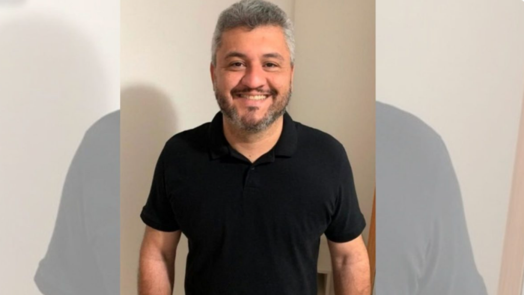 Fotografia de Leopoldo de Barros Teixeira, homem de pele clara, cabelos grisalhos curtos e barba com cavanhaque levemente grisalho, a sorrir para a câmara. Veste uma t-shirt polo preta de mangas curtas e está posicionado no centro da imagem, contra um fundo neutro de tom claro.