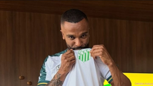Cantor Alexandre Pires segura a camisa de futebol do clube, com o escudo verde e branco visível, em foto interna com luz quente ao fundo.