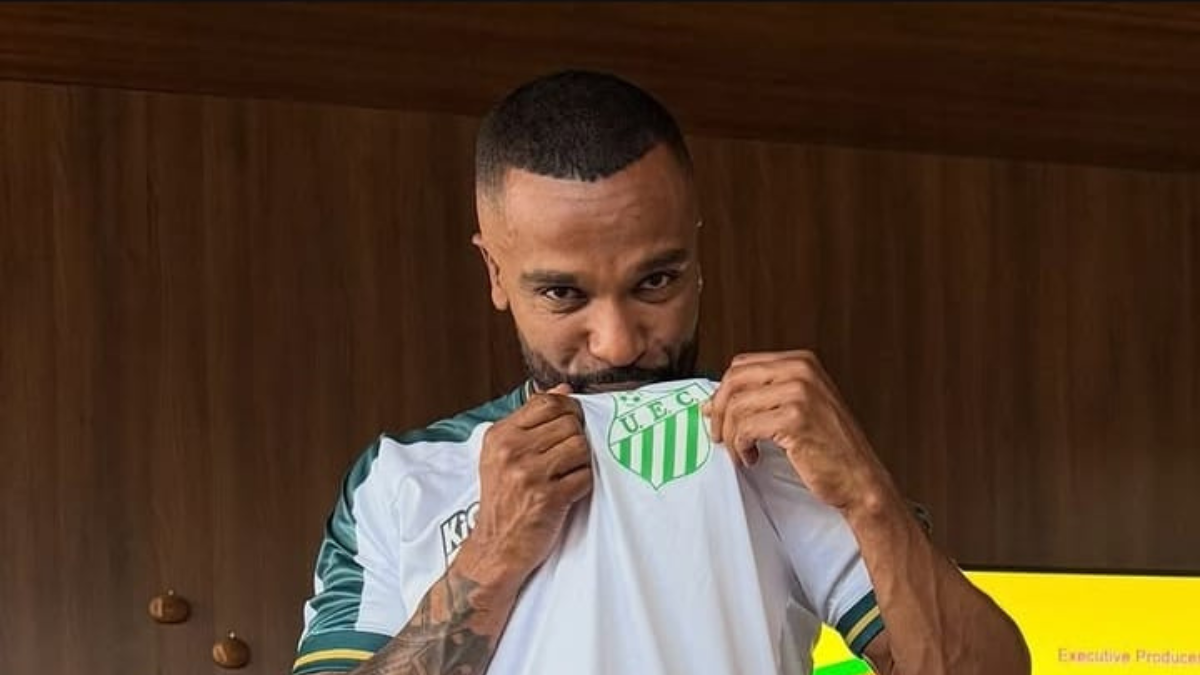 Cantor Alexandre Pires segura a camisa de futebol do clube, com o escudo verde e branco visível, em foto interna com luz quente ao fundo.