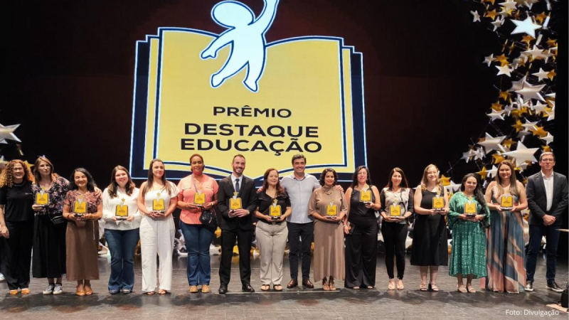 Grupo de homenageados no palco do Teatro Municipal de Indaiatuba, Estado de São Paulo, durante o evento "Prêmio Destaque Educação". As pessoas estão alinhadas, sorrindo e segurando troféus amarelos. Ao fundo, um grande painel exibe o logótipo do prémio e o nome do município, acompanhado por uma decoração suspensa com estrelas prateadas e douradas.