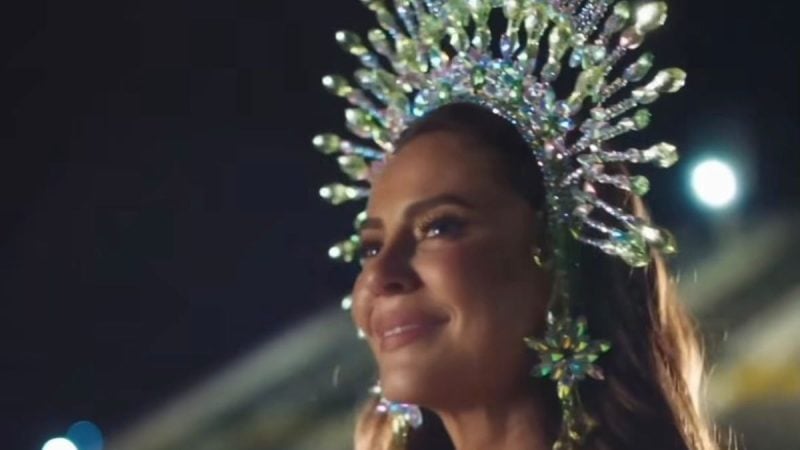 Mulher sorrindo em ambiente noturno, com maquiagem e cabelo presos, usando um adorno luminoso e brilhante em formato de árvore acima da cabeça enquanto aparece com iluminação de festa ao fundo.
