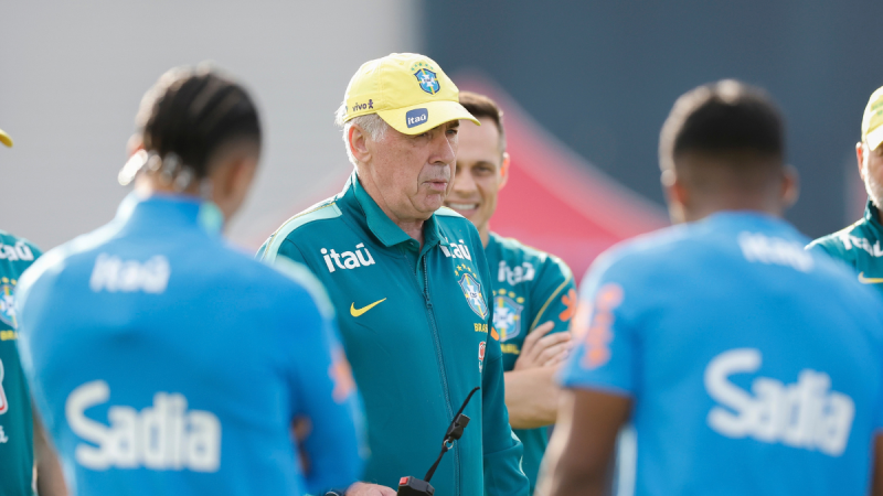Ancelotti à beira do campo durante treino ou jogo da seleção, observando atletas com camisa verde e mãos na beira do rosto.