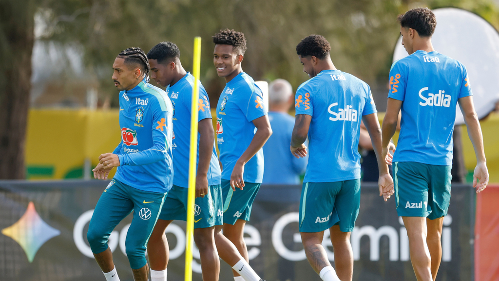 Jogadores da Seleção Brasileira treinando em campo, usando uniforme azul da equipe e camiseta com patrocinadores, durante atividade com bola e cone de treino.