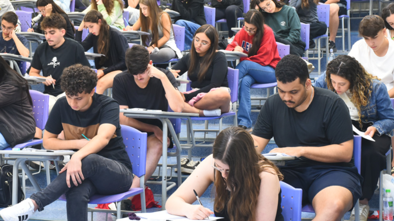 Vestibulandos sentados em salas de aula, fazendo o vestibular da Unicamp com mesas e cadeiras azuis, com materiais sobre as carteiras.