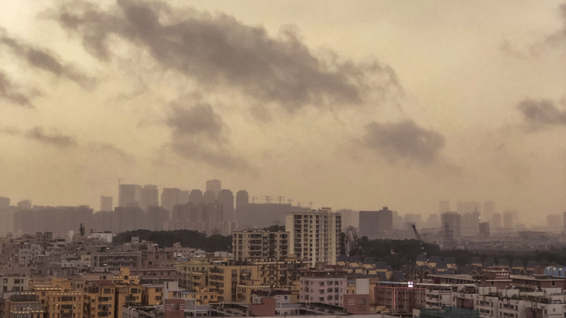Vista panorâmica de uma cidade ao entardecer, com prédios residenciais e comerciais ao fundo, nuvens escuras e céu amarelado iluminado pelo pôr do sol