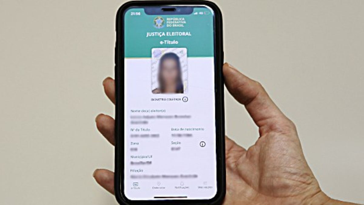 Celular na mão exibindo o app e-Título com a tela da Justiça Eleitoral e comprovante de consulta certificada, em fundo claro.