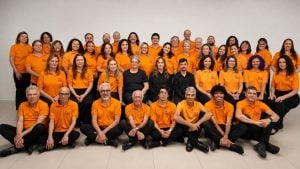 Foto de grupo com colaboradores e líderes vestindo camisetas laranjas, em um ambiente interno, sentados e em pé, organizados para registro institucional. Coral Calixto se apresenta em Santos