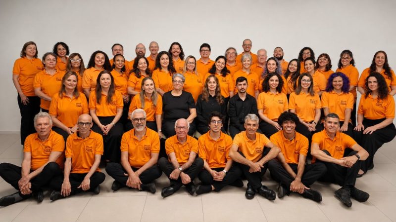 Foto de grupo com colaboradores e líderes vestindo camisetas laranjas, em um ambiente interno, sentados e em pé, organizados para registro institucional. Coral Calixto se apresenta em Santos