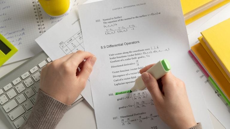 Pessoa estudando cálculo diferencial em uma mesa com laptop e materiais de escritório, segurando um livro com fórmulas e exercícios matemáticos.