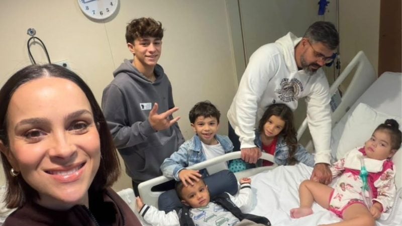 Foto em ambiente hospitalar Leticia Cazarre sorrindo, enquanto os filhos e Juliano Cazarre aparecem ao fundo, sentados em macas e com equipamentos médicos, durante uma visita médica