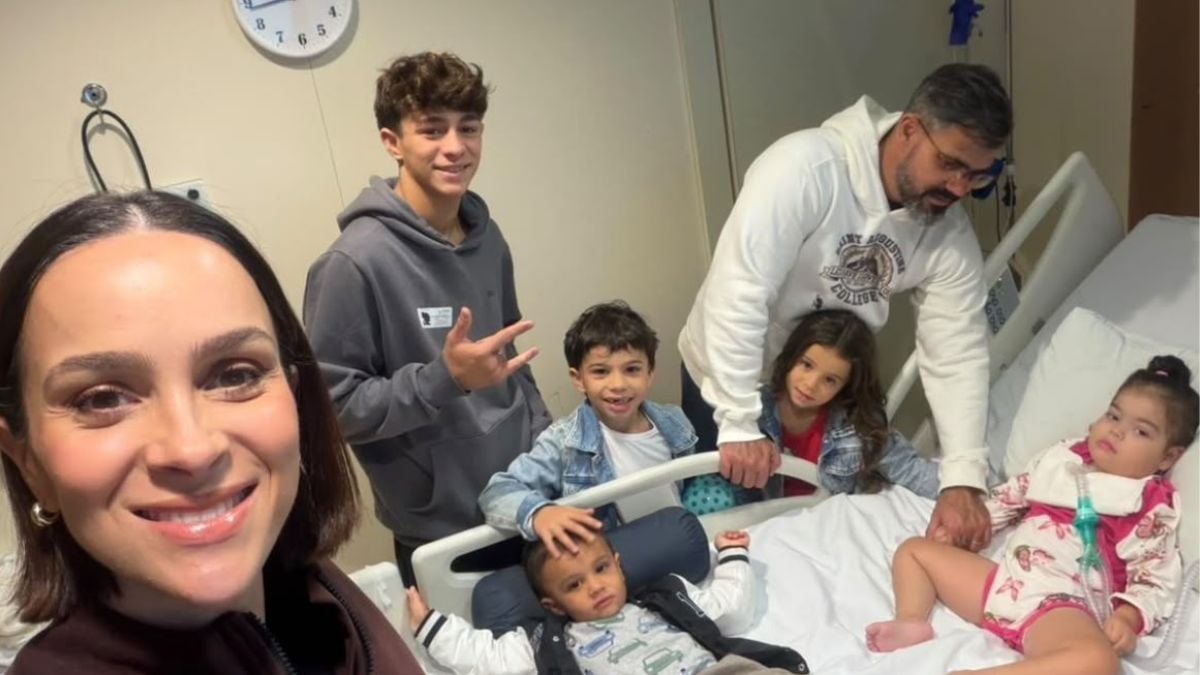 Foto em ambiente hospitalar Leticia Cazarre sorrindo, enquanto os filhos e Juliano Cazarre aparecem ao fundo, sentados em macas e com equipamentos médicos, durante uma visita médica