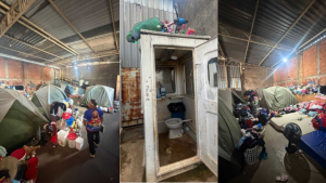 Interior de um abrigo com tendas e pessoas em situação de acolhimento, ao lado de uma instalação sanitária com vaso e pia em ambiente de galpão
