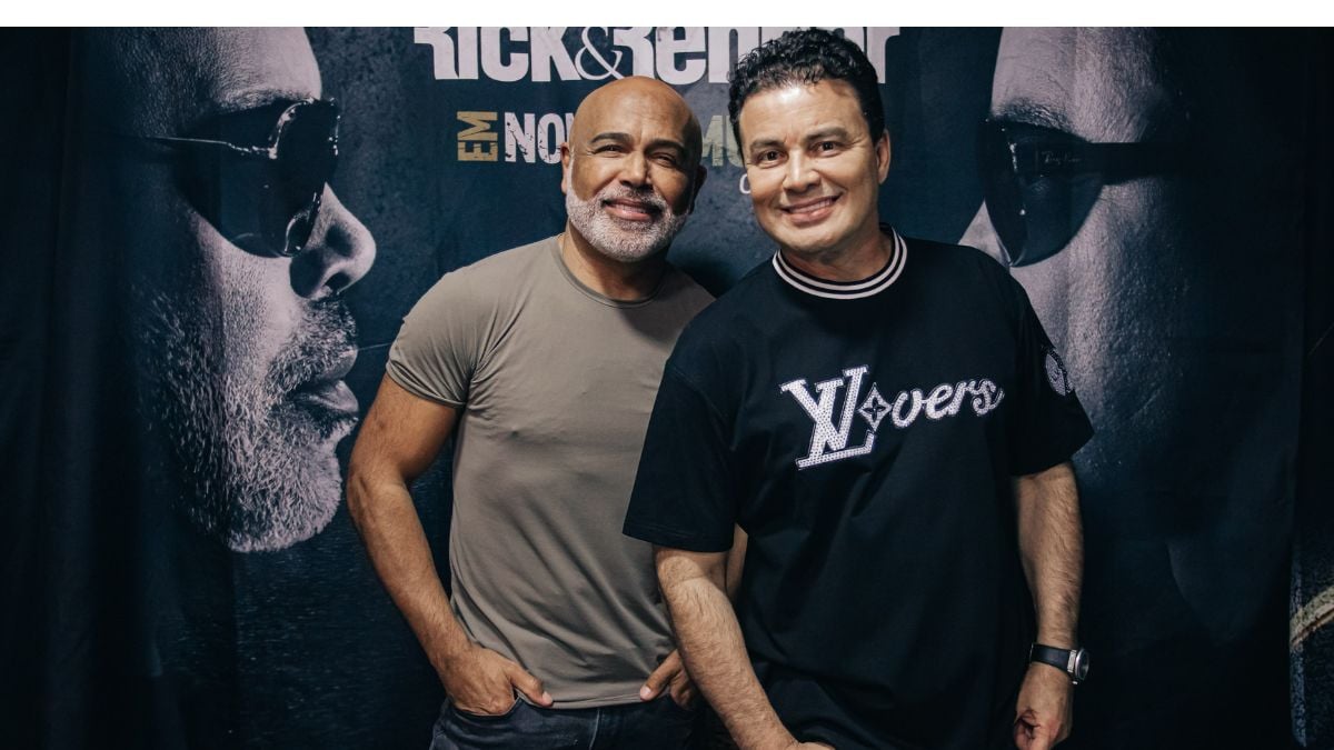 Rick & Renner posam lado a lado em frente a um cartaz com grande destaque para rostos e texto em estilo promocional, em foto de evento.