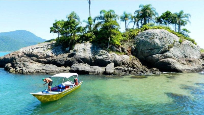Barco a vela pequeno no mar de águas verdes próximo a Ilha do guaraú, em Peruíbe, rochosa com vegetação tropical e montanha ao fundo, em cenário de passeio.
