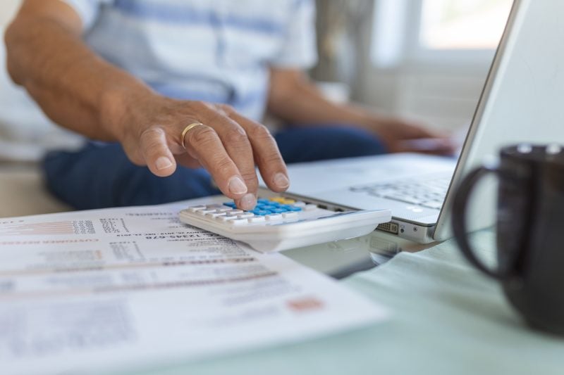 Pessoa usando calculadora azul e analisando documentos financeiros sobre uma mesa, enquanto trabalha com laptop em ambiente de escritório