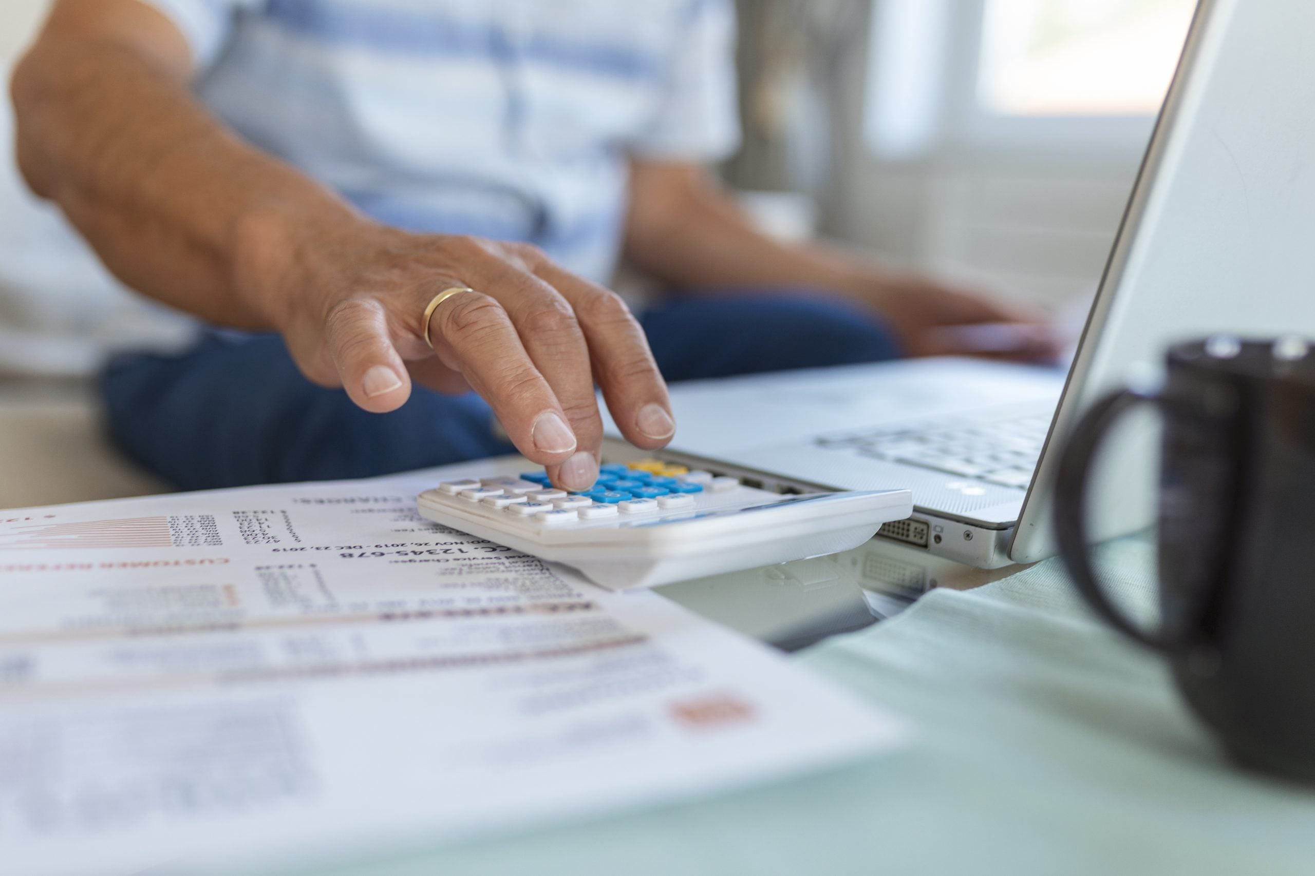 Pessoa usando calculadora azul e analisando documentos financeiros sobre uma mesa, enquanto trabalha com laptop em ambiente de escritório