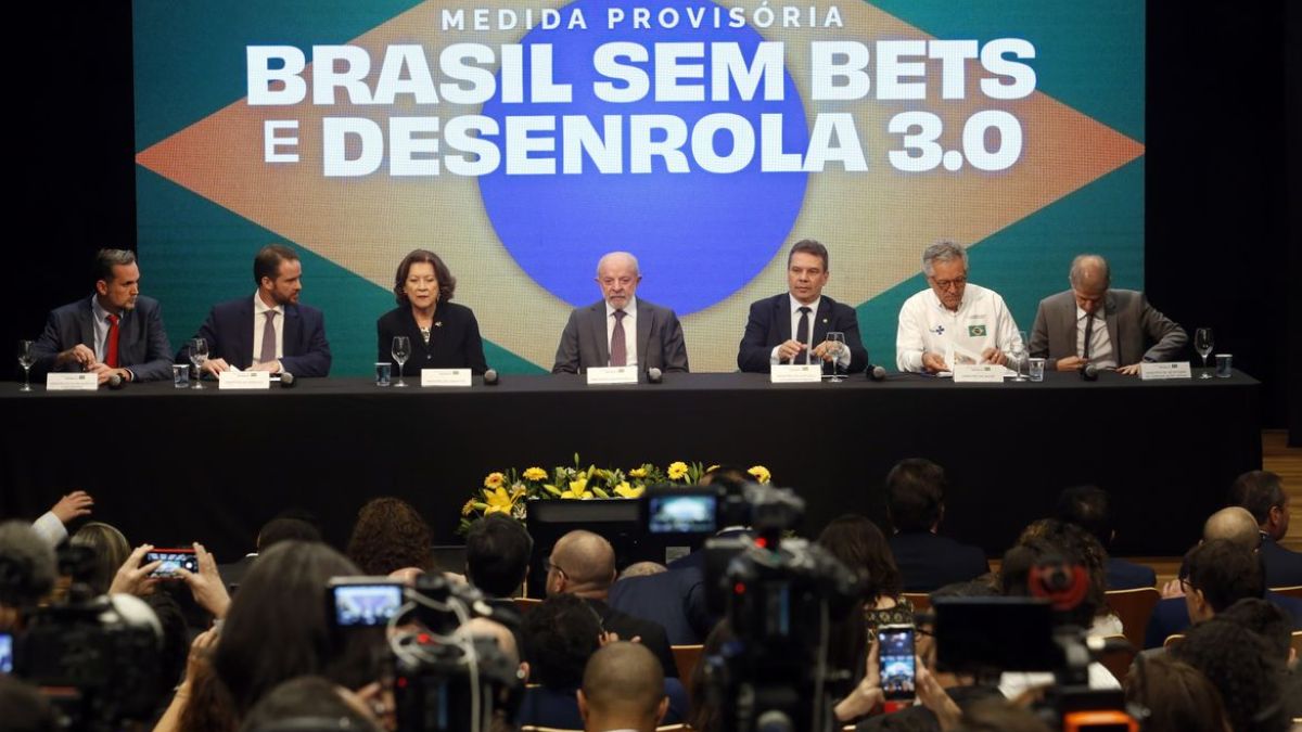 Bancada com o PresidenteLula ao lado de ministros em anuncio do novo Desenrola. Ao fundo, um painel com a bandeira do Brasil escrito "Brasil sem Bets e Desenrola 3.0