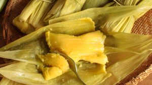 Pamonha aberta com garfo cortando a comida ao meio. Ao redor várias pamonhas fechadas na folha do milho