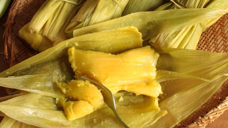Pamonha aberta com garfo cortando a comida ao meio. Ao redor várias pamonhas fechadas na folha do milho