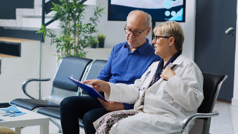 Duas pessoas, um paciente e uma médica, sentados em uma sala de atendimento, analisando documentos em uma prancheta e conversando com seriedade.