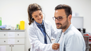 Médica com estetoscópio e jaleco branco observa e orienta um paciente em ambiente clínico, enquanto ele usa óculos, ambos com foco em avaliação de saúde.