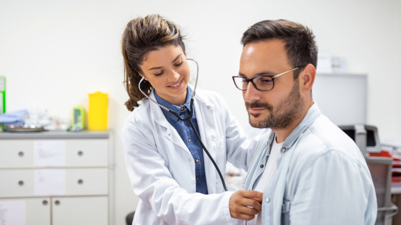 Médica com estetoscópio e jaleco branco observa e orienta um paciente em ambiente clínico, enquanto ele usa óculos, ambos com foco em avaliação de saúde.