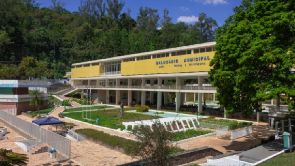 Prédio público municipal em meio a área verde, com gramado, jardins e caminhos pavimentados em frente à construção. Fachada amarela e janelas amplas sob céu azul.
