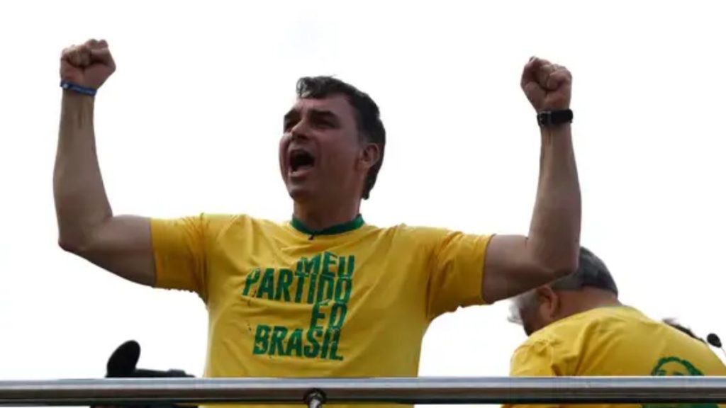 Flávio Bolsonaro comemora com os braços para cima, está de camiseta amarela e com a frase em verde "MEU PARTIDO É O BRASIL"