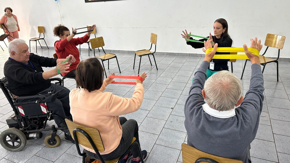 Aula de atividade física para idosos em grupo, com cadeiras e participantes em cadeiras de rodas fazendo exercícios com faixas coloridas orientados por uma instrutora ao fundo.