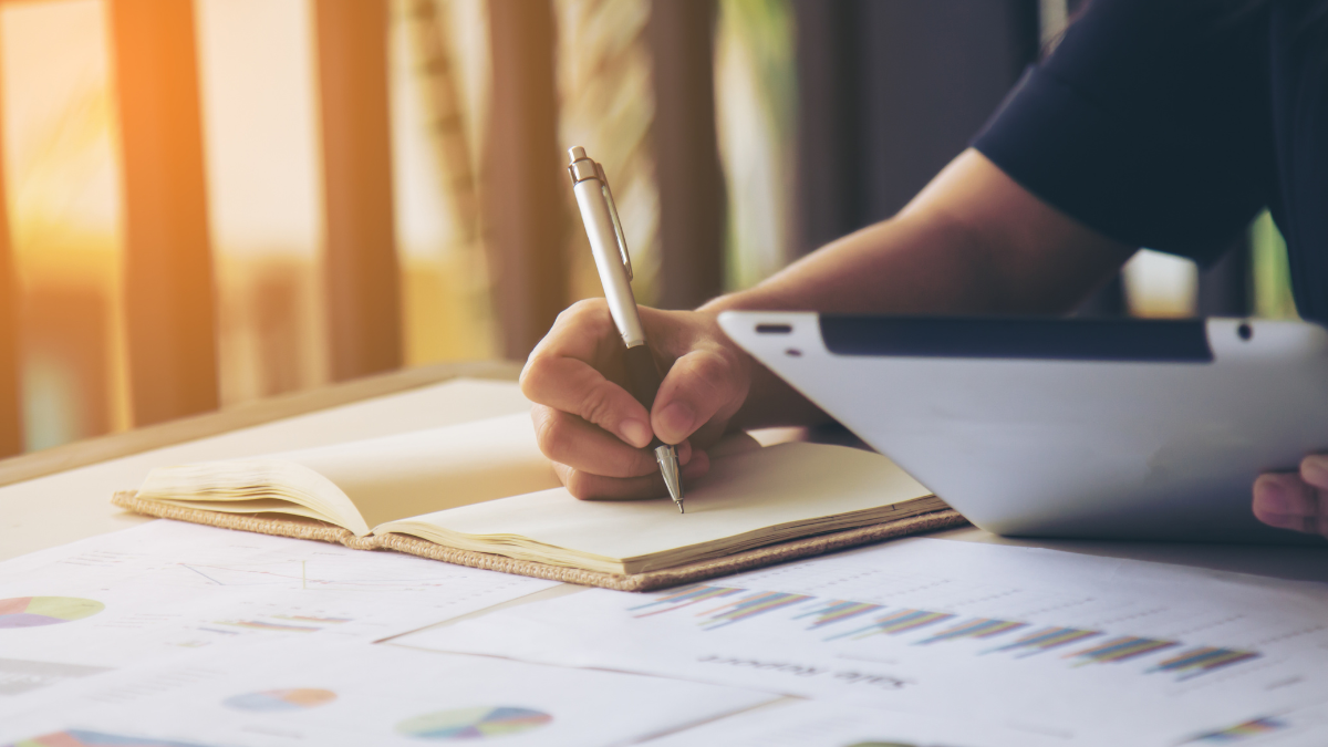 Pessoa segurando uma caneta escreve em um papel sobre uma mesa com gráficos e documentos, enquanto usa um notebook ao lado, em ambiente iluminado ao pôr do sol