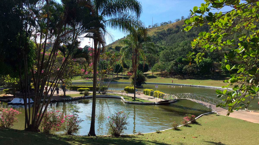 Paisagem de jardim em área rural com lago central, trilhas e passarela de madeira, palmeiras e vegetação verde, flores em canteiros e colinas ao fundo sob céu azul