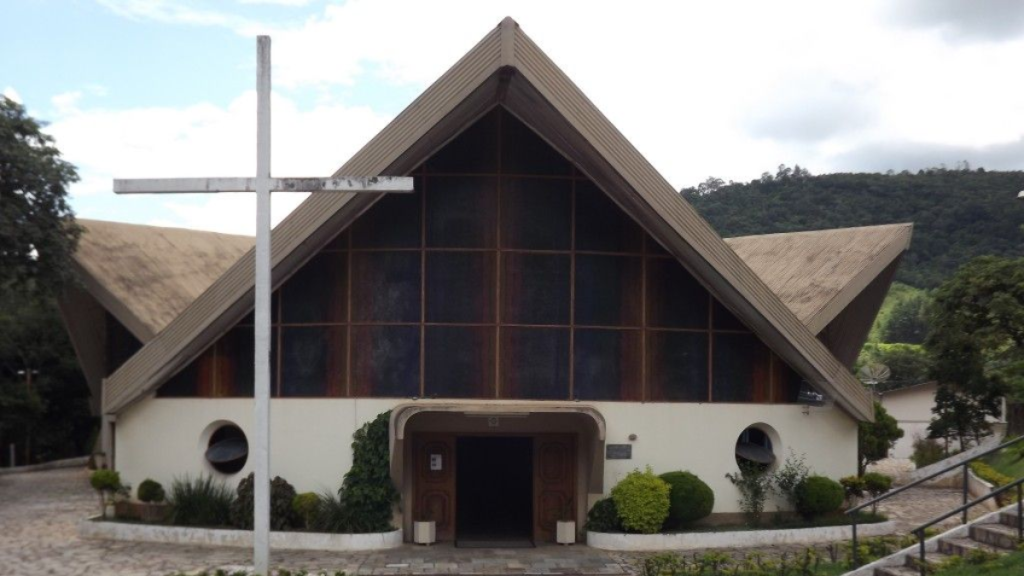 Fachada de uma igreja moderna de madeira e vidro, com cruz branca na frente, janelas em grade, vegetação ao redor e entrada central com portas de madeira em meio a área verde e colinas.