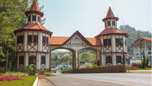Entrada de um complexo com portões em arco e duas torres de madeira e telhado vermelho, em ambiente verde e cercado por jardins, com caminho de acesso à frente.