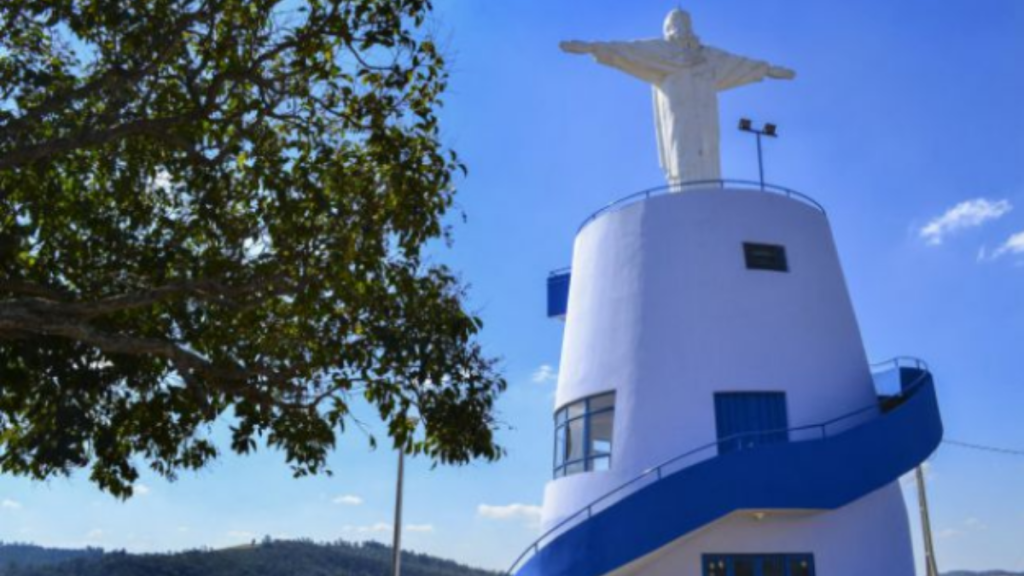 Imagem de um farol branco e azul com uma estátua no topo, visto contra o céu azul. Ao lado há árvores verdes e paisagem ao fundo.