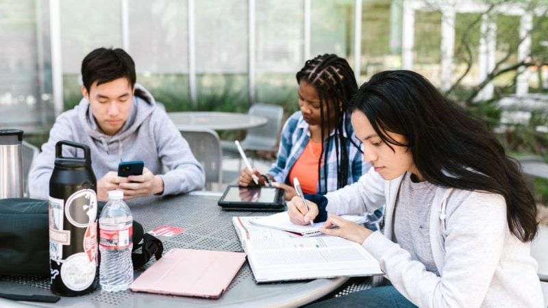 Estudantes em uma biblioteca, com cadernos e tablets, fazendo anotações e respondendo questões, em um contexto de preparação para o Enem e uso da nota no Sisu, Prouni e Fies.