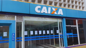 Fachada de uma agência bancária da Caixa Económica Federal. Na parte superior, destaca-se o letreiro azul com o logótipo da "CAIXA" em letras brancas, sobressaindo a letra "X" em tons de laranja e branco. Abaixo do letreiro, a entrada possui portas e janelas de vidro transparente com avisos colados, além de faixas azuis sinalizadas com o símbolo internacional de acessibilidade.