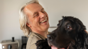 Mulher idosa sorrindo ao lado de um cachorro preto em ambiente interno, com expressão de carinho e bem-estar, perto de equipamentos domésticos ao fundo.