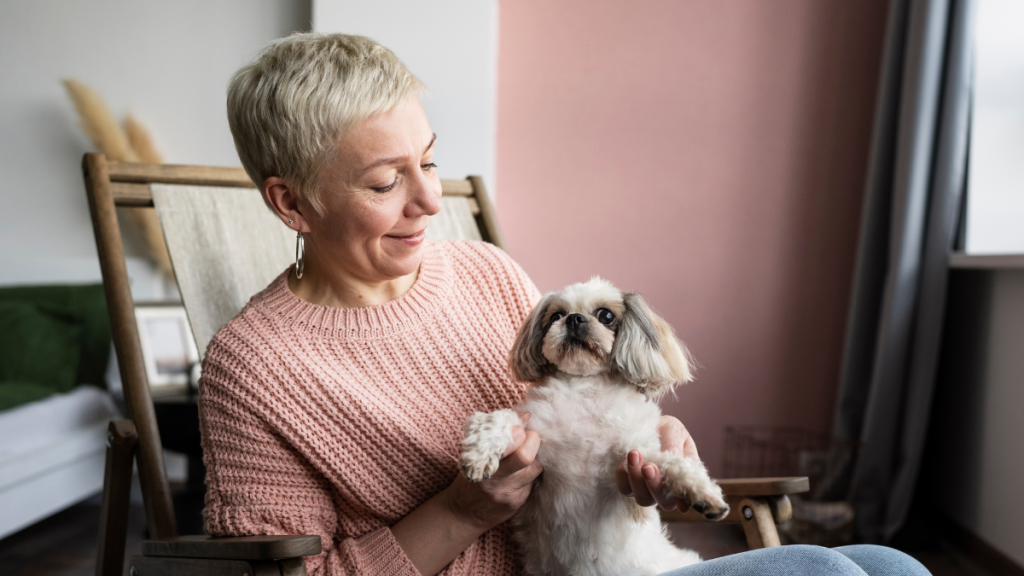 Mulher idosa sorridente abraçando e segurando um cachorrinho pequeno e peludo no colo, em um ambiente interno acolhedor, com luz natural suave.