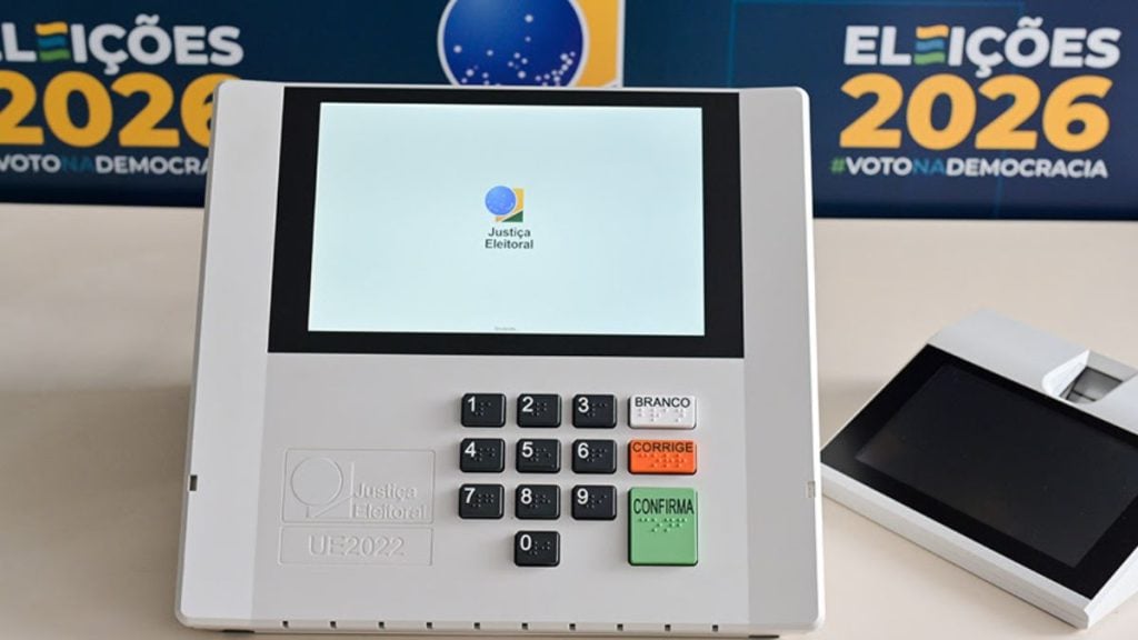 Urna eletrônica com tela exibindo o logo da Justiça Eleitoral e teclado numérico, com painel de confirmar, corrigir e opções de voto branco ao lado de outros equipamentos de votação.