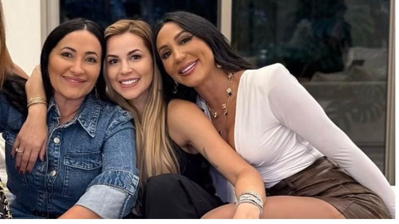 Deolane Bezerra, Dayanne Bezerra e mãe sentadas juntas posando para uma foto, com roupas casuais e sorrindo ao ar livre, em um ambiente com fundo desfocado.