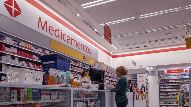Interior de uma farmácia com prateleiras de medicamentos e caixas organizadas, faixa de sinalização escrito “Medicamentos” e atendimento ao balcão, com cliente ao lado do guichê.