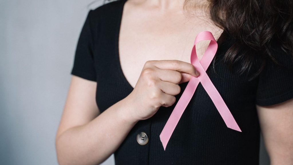 Câncer de mama: mulher usando fita rosa de conscientização, apontando para o laço no peito em campanha sobre prevenção e pós-tratamento.