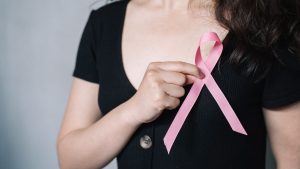 Câncer de mama: mulher usando fita rosa de conscientização, apontando para o laço no peito em campanha sobre prevenção e pós-tratamento.