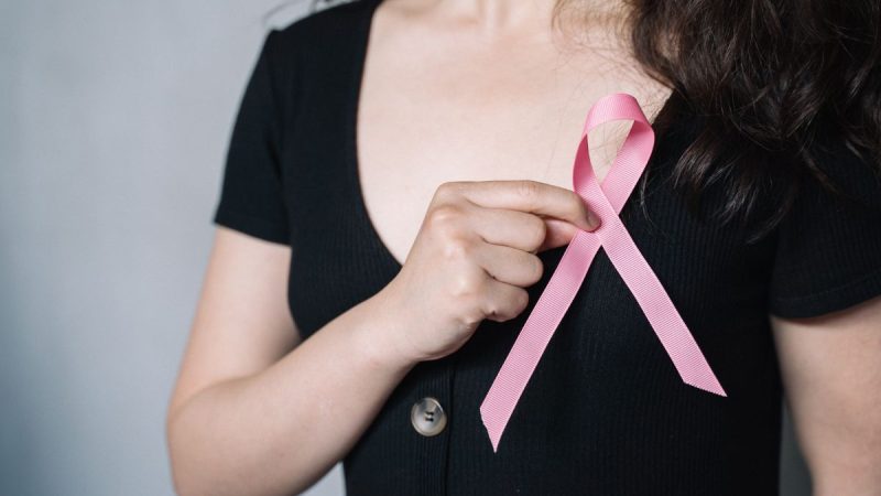 Câncer de mama: mulher usando fita rosa de conscientização, apontando para o laço no peito em campanha sobre prevenção e pós-tratamento.